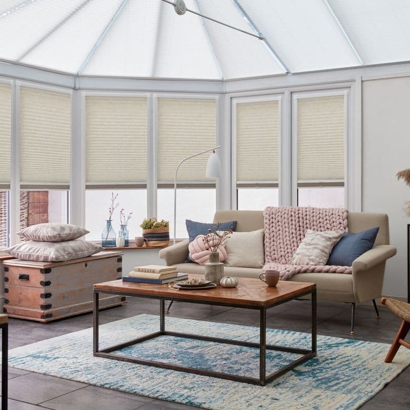 Pleated honeycomb blinds covering conservatory’s tall, multi-panel windows, lowered to mid-height and filtering soft daylight; beige blinds frame a cozy sunroom with sofa, knitted throw, wooden coffee table and rug.