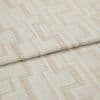 Beige woven fabric with light-brown geometric square pattern (curtain/upholstery fabric) folded over itself, revealing textured weave; close-up studio product view under soft, neutral lighting.
