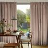 Floor-to-ceiling pleated beige-pink curtains, drawn partially aside to reveal a glass door; curtains filtering daylight. Dining table with pancakes in a warmly lit kitchen opening onto a green garden.