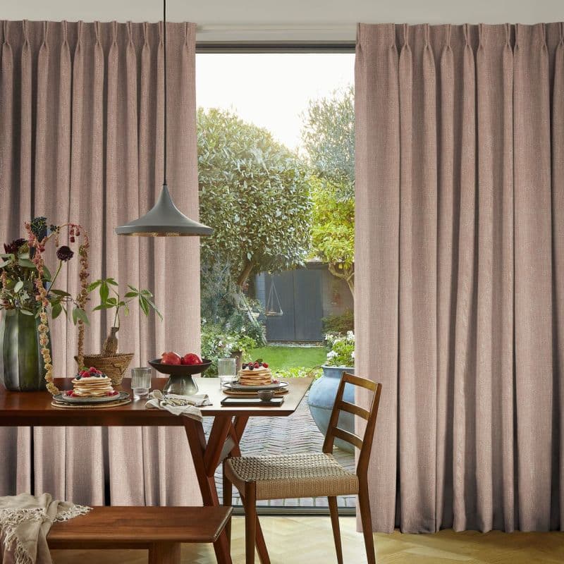 Floor-to-ceiling pleated beige-pink curtains, drawn partially aside to reveal a glass door; curtains filtering daylight. Dining table with pancakes in a warmly lit kitchen opening onto a green garden.