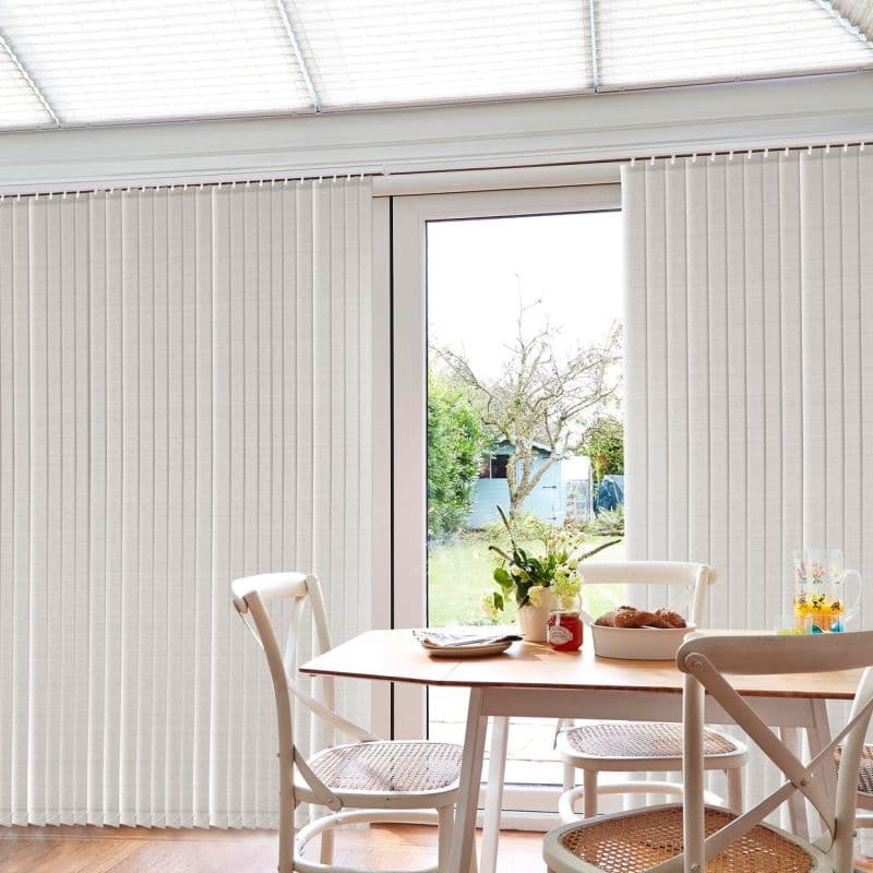 Cream vertical blinds, full-length across a sliding door, parted to sides; partially open and filtering soft daylight; bright dining area with wooden table, chairs, vase of flowers and garden view.