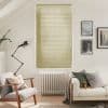 Horizontal beige Venetian blinds covering a tall window, fully closed and softly filtering daylight; living room with armchair, radiator, floor lamp, sofa, cushions and framed wall art in neutral tones.