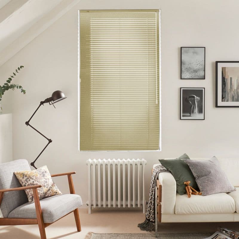 Horizontal beige Venetian blinds covering a tall window, fully closed and softly filtering daylight; living room with armchair, radiator, floor lamp, sofa, cushions and framed wall art in neutral tones.