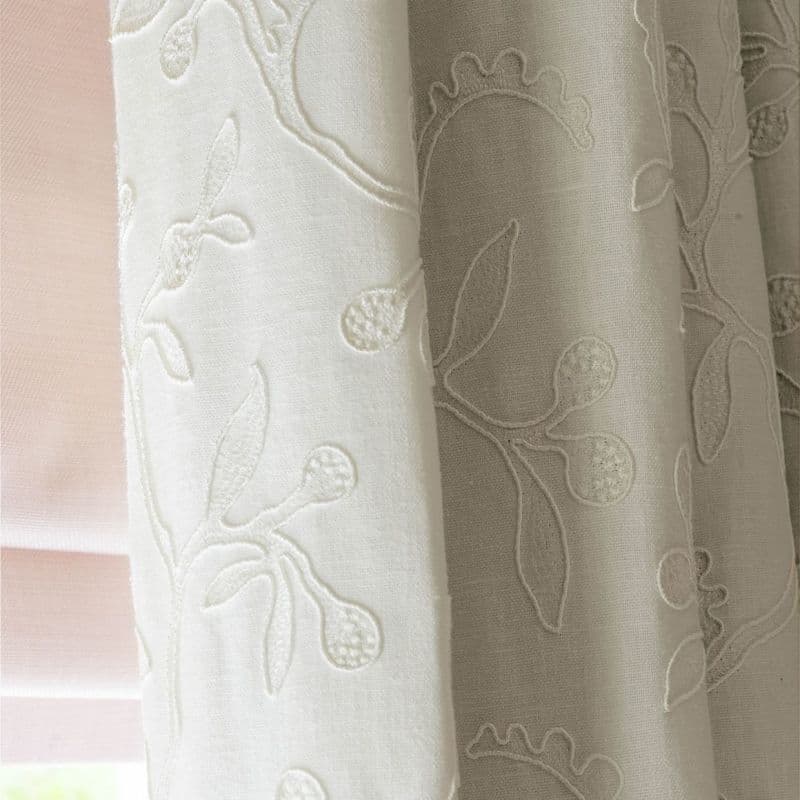 Cream embroidered linen curtains, hanging in soft folds over a pale pink Roman blind, filtering diffused daylight; close-up of an interior window with muted, warm lighting.