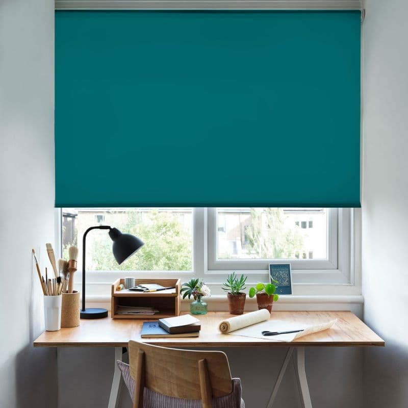 A teal roller blind pulled down fully across a double window, blocking most light; a wooden desk beneath holds a lamp, plants, notebooks, brushes, and daylight through a lower gap.
