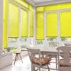 Yellow pleated blinds cover tall French doors and windows, lowered and filtering daylight into a sunlit dining nook with a wooden table, chairs, and a garden view.