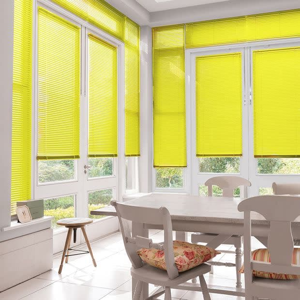 Yellow pleated blinds cover tall French doors and windows, lowered and filtering daylight into a sunlit dining nook with a wooden table, chairs, and a garden view.