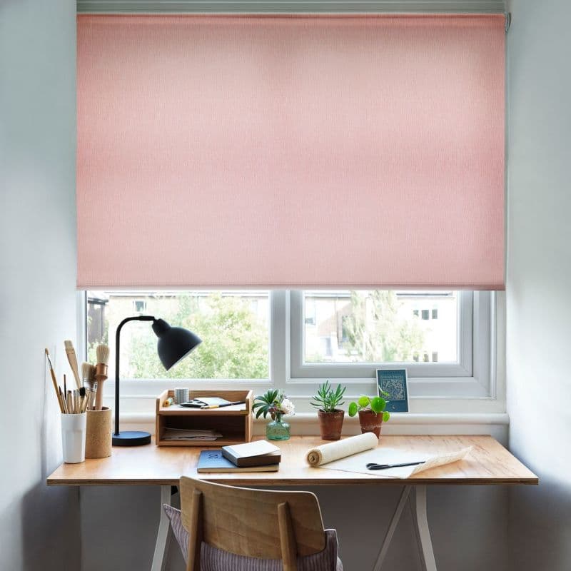 Soft pink textured roller blind, lowered across the window, filtering daylight; tidy wooden desk beneath holds a lamp, paintbrushes, notebooks, small potted plants and chair in a neutral-lit room.