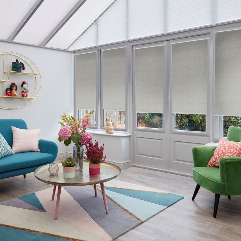 Light grey roller blinds, lowered over tall glazed doors and windows, partially blocking sunlight; conservatory-style living room with teal sofa, green armchair, pastel geometric rug, round coffee table and flowers.
