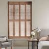 Wooden plantation shutters — four full-height panels with horizontal louvers — cover a recessed window, closed and angling slats to filter daylight in a neutral bedroom with armchair, bed, side table.