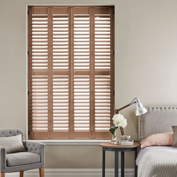 Wooden plantation shutters — four full-height panels with horizontal louvers — cover a recessed window, closed and angling slats to filter daylight in a neutral bedroom with armchair, bed, side table.