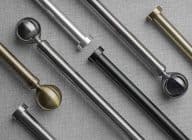 Curtain rods with varied metallic finials (spherical, hexagonal), brass, chrome and black finishes, lie diagonally and overlapped, displayed on gray woven fabric under neutral studio lighting.