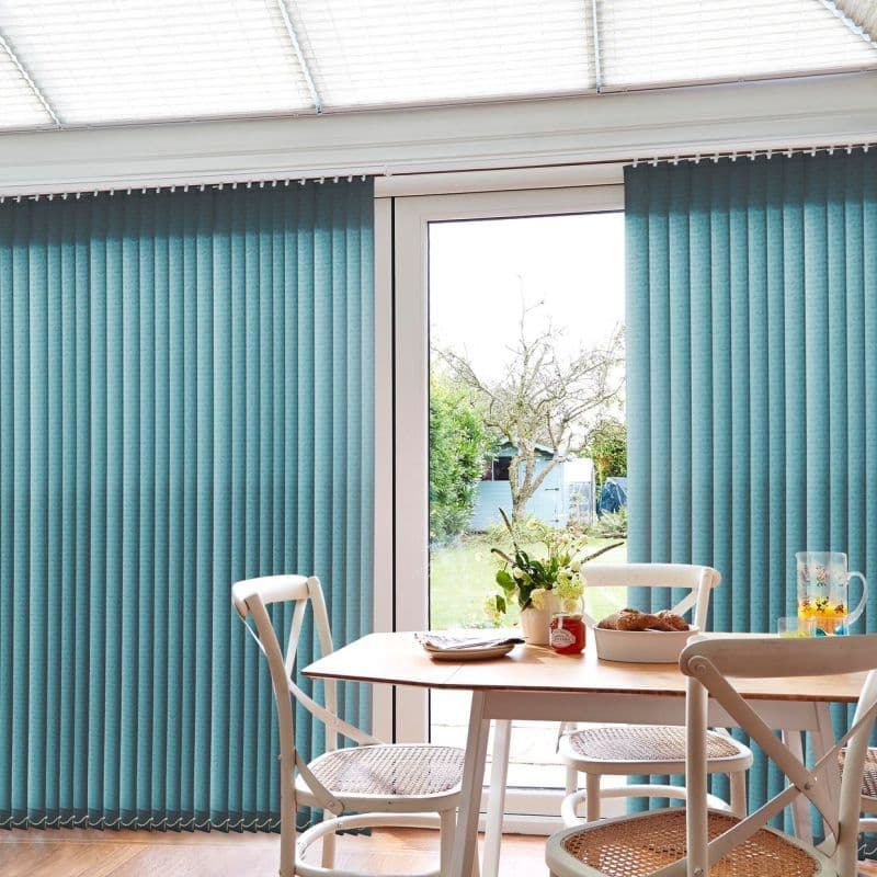 Teal fabric vertical blinds, covering a sliding glass door with slats closed except central gap; filtering daylight into dining nook with wooden table, chairs, vase and garden view.