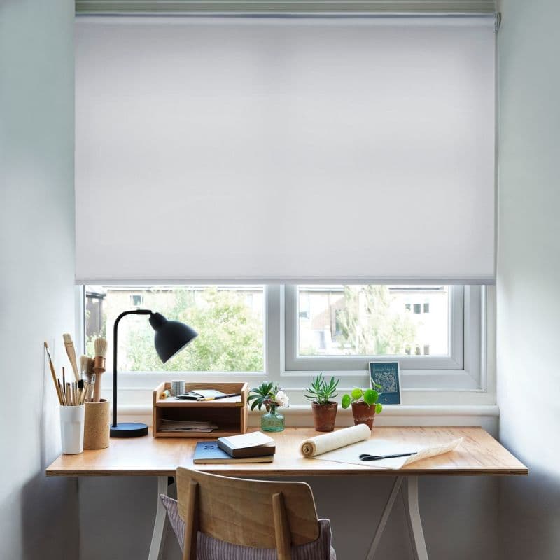 White fabric roller blind, lowered, softly filtering daylight over a double‑pane window; wooden desk below holds a lamp, art brushes, notebooks, potted succulents and a chair.