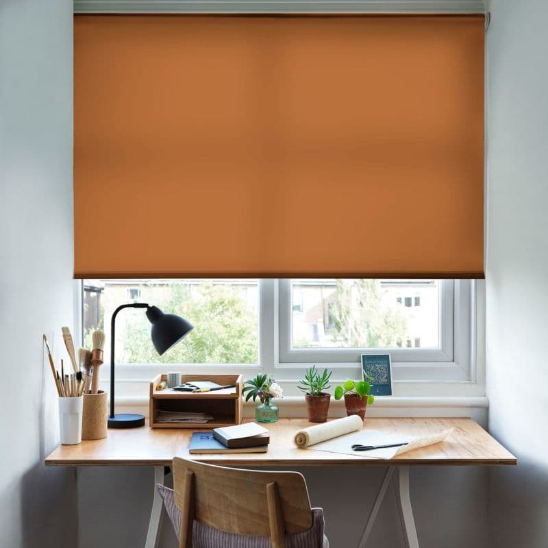 Orange roller blind lowered almost fully across a double-pane window, filtering daylight; a wooden desk beneath holds a black lamp, brushes, notebooks and potted succulents in a bright study nook.