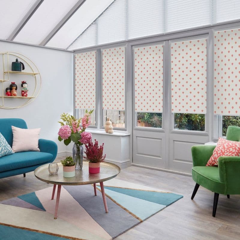 Patterned roller blinds, white with small pink teardrop motifs, lowered over tall conservatory windows and doors, filtering daylight; room shows angled glass roof, colorful sofas, rug, coffee table and plants.