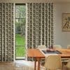 Geometric-print, full-length curtains with a linked-chain motif, hung floor-to-ceiling and parted to frame a narrow central window, filtering daylight into a mid-century dining room with wooden table and garden view.