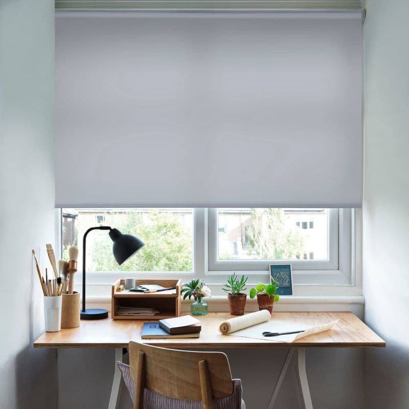 White roller blind lowered across a double window, filtering daylight; below, a wooden desk with lamp, artbrushes, notebooks and small potted plants, overlooking leafy buildings outside.