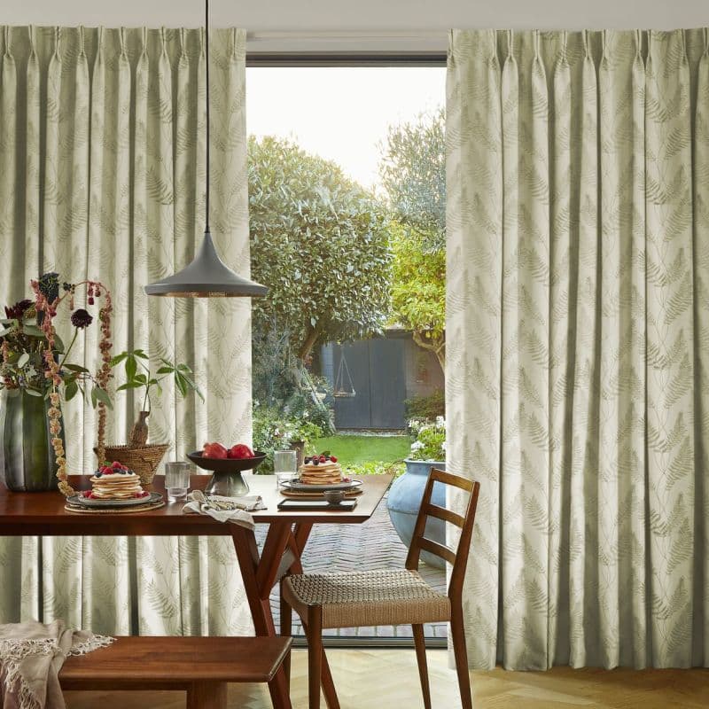 Pleated cream curtains with fern print, hung beside a sliding door, parted to frame and softly filter daylight, overlooking a sunlit backyard from a breakfast dining table.