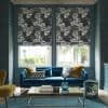 Floral Roman blinds over two tall windows, lowered and patterned with large monochrome roses, filtering daylight into a teal living room with a blue velvet sofa and glass coffee table.
SCARPA