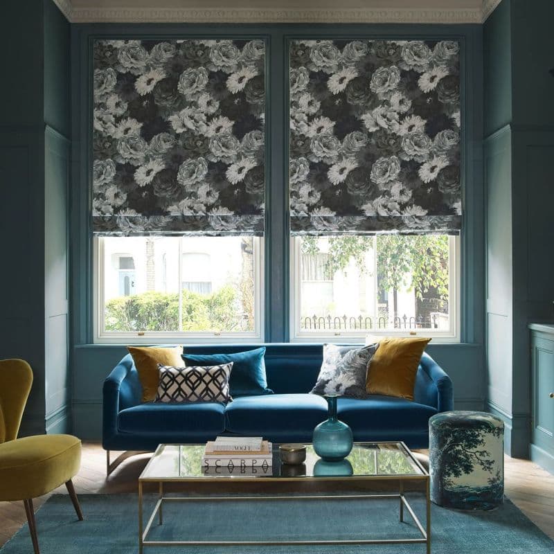 Floral Roman blinds over two tall windows, lowered and patterned with large monochrome roses, filtering daylight into a teal living room with a blue velvet sofa and glass coffee table.
SCARPA