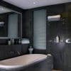 Narrow vertical venetian blinds covering a tall bathroom window, closed and filtering soft daylight; dark slate-tiled walls, freestanding tub, mirrored shelf, towels and toiletries nearby.