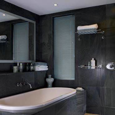 Narrow vertical venetian blinds covering a tall bathroom window, closed and filtering soft daylight; dark slate-tiled walls, freestanding tub, mirrored shelf, towels and toiletries nearby.