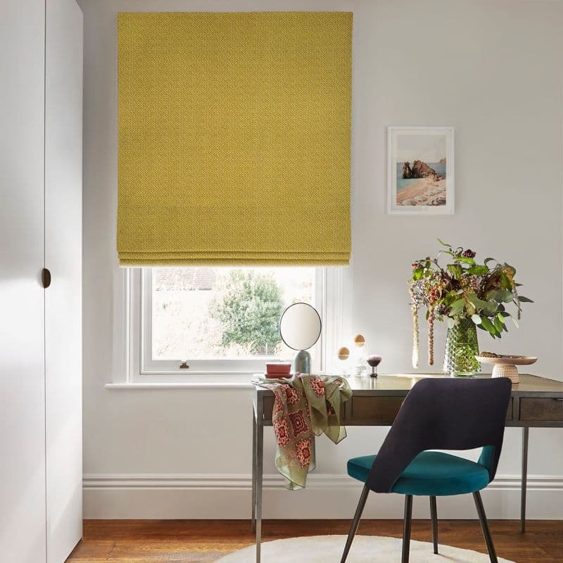 A mustard yellow Roman blind, fully lowered with subtle textured weave, filtering soft daylight from a sash window above a wooden desk with chair, vase of flowers and coastal print.