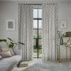 Patterned drapery panels, light grey with a geometric motif, hung on a rod and parted; filtering soft daylight through tall black-framed French doors into a living room overlooking a garden.