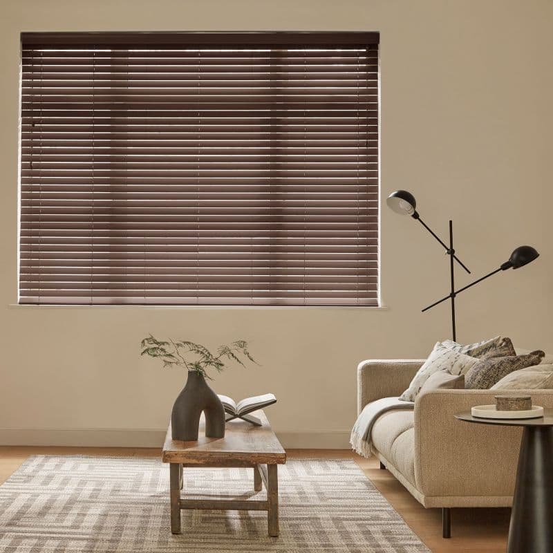 Brown horizontal Venetian blinds covering a large window, fully closed and filtering soft daylight through narrow slats; a neutral living room with beige sofa, floor lamp, wooden coffee table and rug.