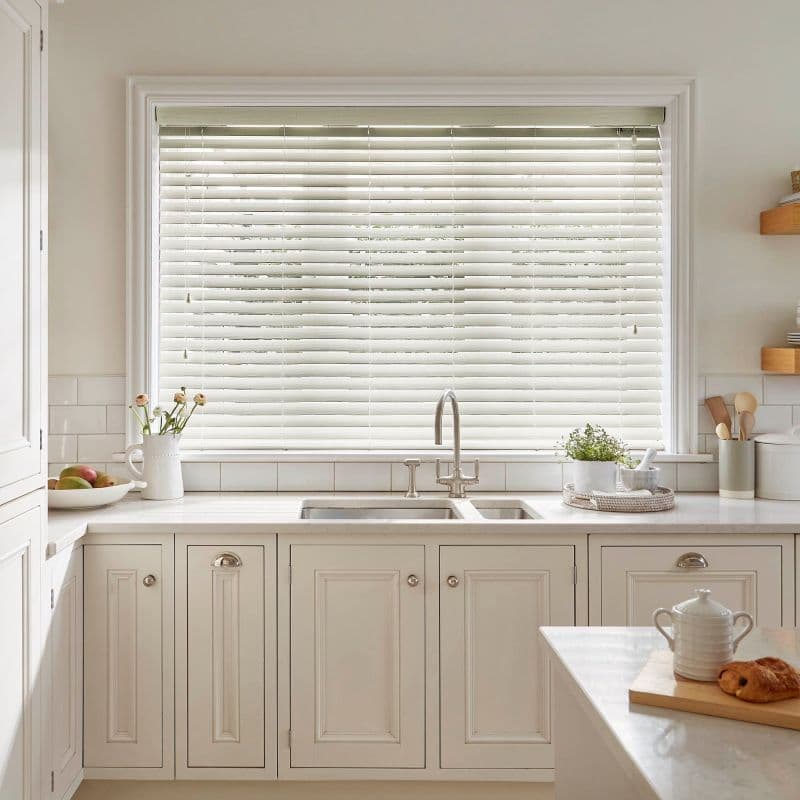 White horizontal Venetian blinds cover a large window, slats mostly closed and softly filtering daylight; above a farmhouse sink in a light, cream kitchen with marble counters, plants, and utensils.