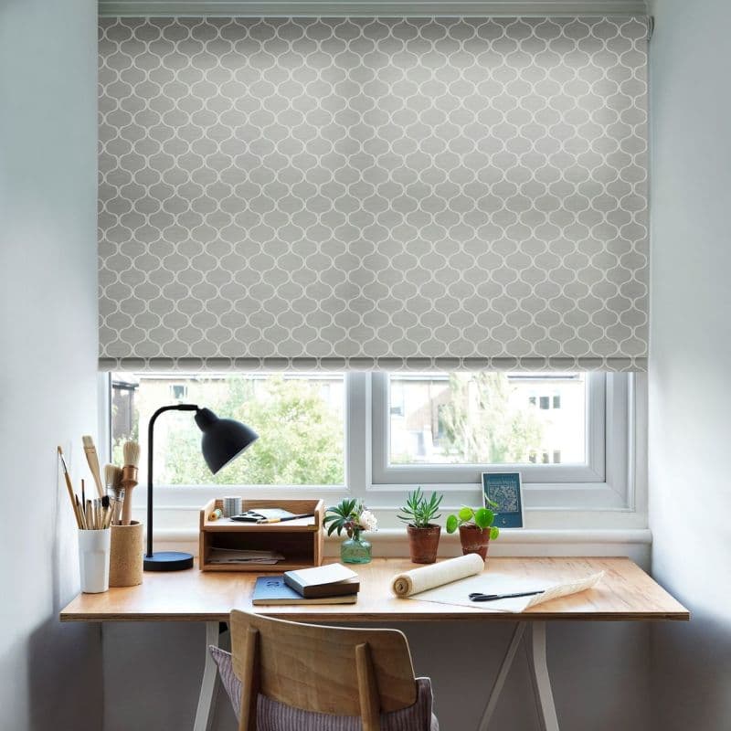 A gray roller blind with white interlocking quatrefoil pattern, fully lowered and filtering daylight over a compact wooden desk with lamp, succulents, art supplies and chair in a bright room.