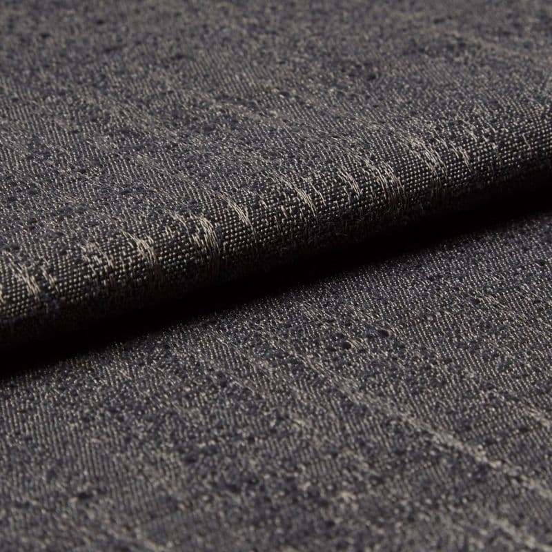 Dark gray woven fabric with slubs and subtle metallic threads; folded into a layered edge; tight macro close-up on a softly lit surface, neutral background suggesting an upholstery sample.