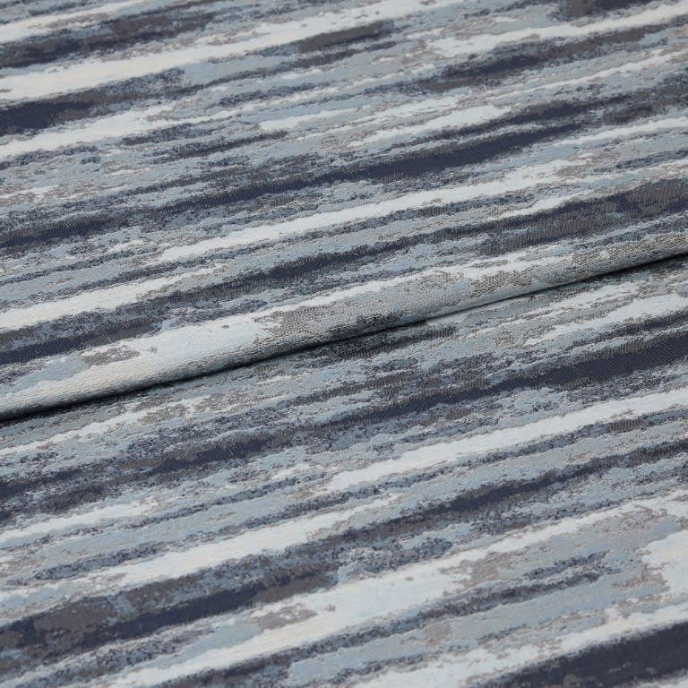 Folded jacquard curtain fabric with horizontal blue-gray, navy, and white striations; draped over itself, revealing woven texture and subtle sheen in a close-up, softly lit studio view.