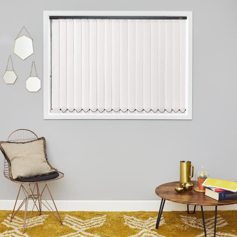 White vertical blinds covering a rectangular window; closed slats with bead chain at bottom, filtering light, in a grey-walled living area with a wire chair, mustard rug and wooden coffee table.