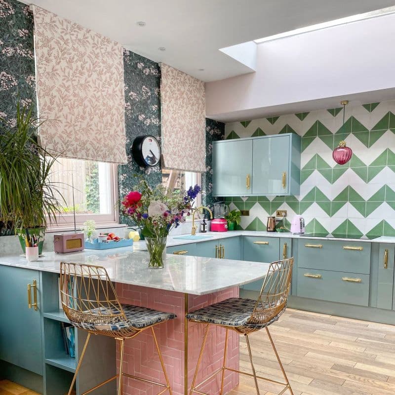 Three fabric Roman blinds, pale pink floral panels lowered over a wide window, filtering daylight; kitchen with pastel teal cabinets, pink-tiled island, marble countertop, green chevron wall tiles, skylight.