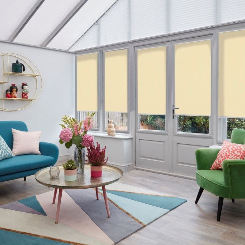 Cream roller blinds lowered over tall French doors and windows, covering panels and leaving clear lower panes, filtering daylight into a bright conservatory with teal sofa, green armchair, pastel rug.