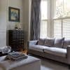 Floor-to-ceiling pleated gray curtains alongside white café-style louvered shutters covering a bay window, partially closed and filtering bright daylight; modern living room with gray sofa, ottoman, dark cabinet.