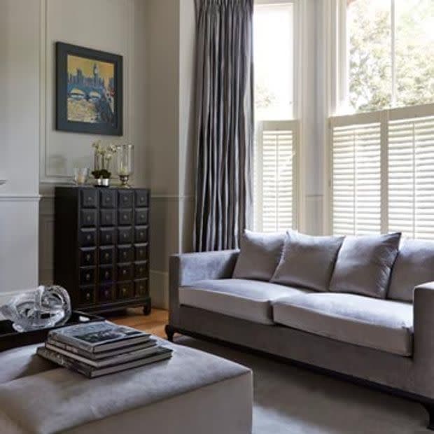 Floor-to-ceiling pleated gray curtains alongside white café-style louvered shutters covering a bay window, partially closed and filtering bright daylight; modern living room with gray sofa, ottoman, dark cabinet.