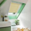 A pleated yellow skylight blind, mounted inside the frame, is partially lowered, filtering daylight over a sloped bathroom with green-painted trim, white herringbone tiles, sink, tub, and plant.