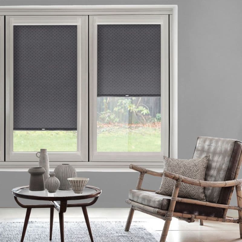 Three grey patterned roller blinds cover a triple window — the right blind partially raised, filtering soft daylight into a neutral living room with a rattan armchair and small decorative table.