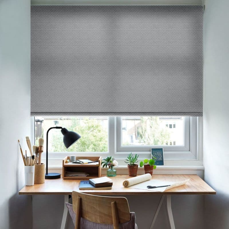 Grey patterned roller blind lowered halfway across double window, filtering daylight; wooden desk beneath holds black lamp, art supplies, potted plants and notebooks in a bright home workspace.