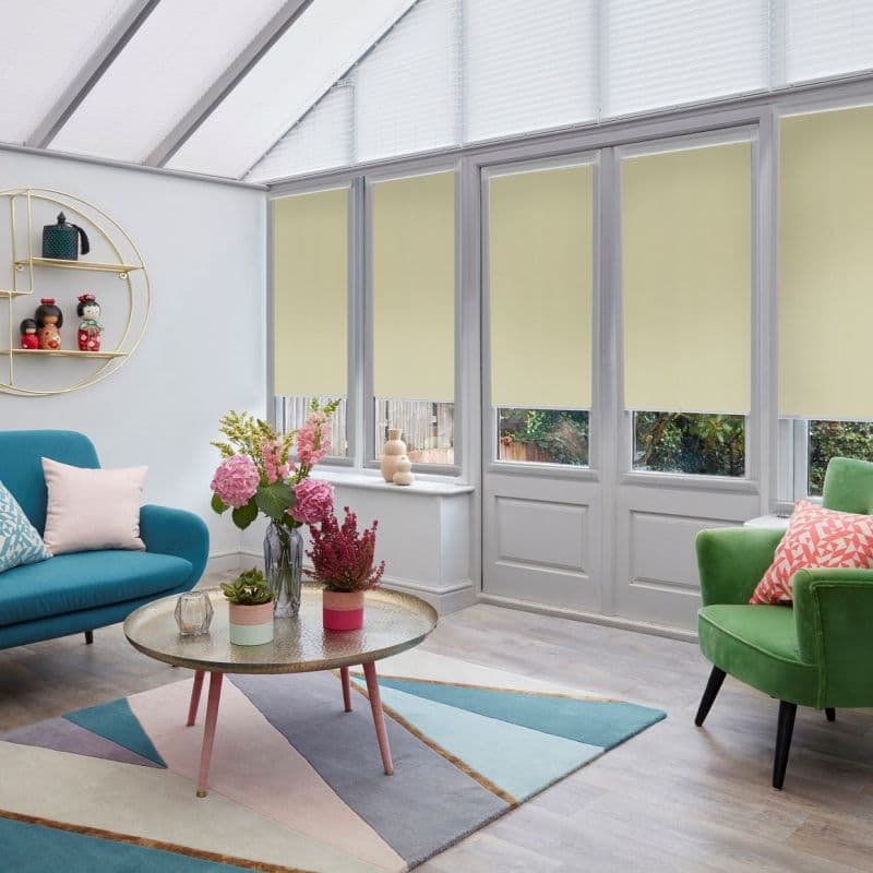 Pale yellow roller blinds across tall glazed doors and upper windows, partially lowered, filtering daylight; in a bright conservatory-style living room with colorful sofas, round coffee table, flowers.