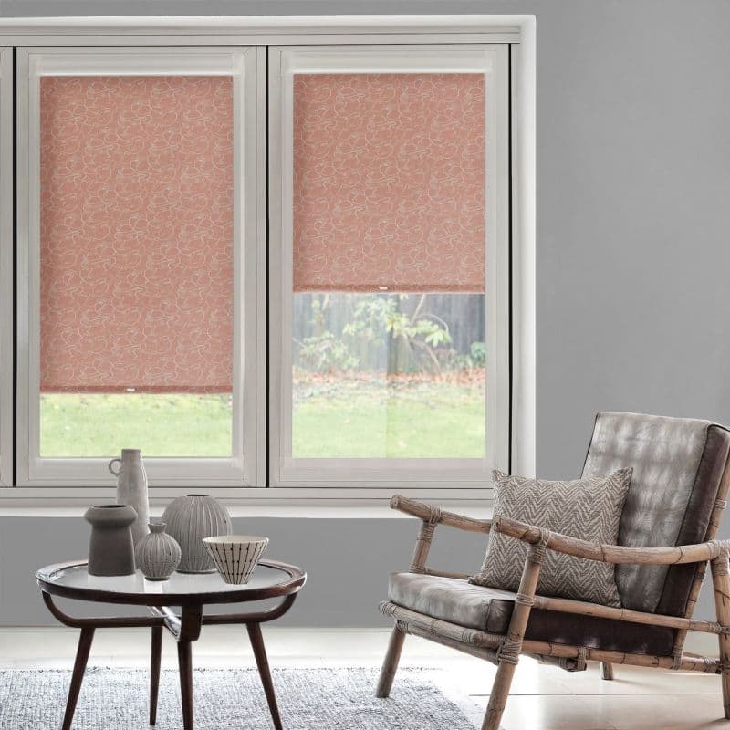 Two pink floral roller blinds in a white-framed double window—left fully lowered, right partially lowered filtering daylight; living room with gray walls, rattan armchair, small table with ceramics, garden view.
