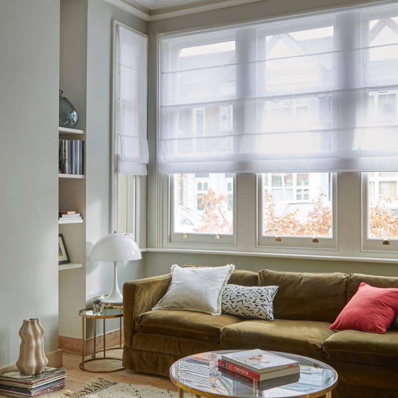 Sheer white Roman blinds lowered across a bay of windows, softly diffusing daylight; a living room with an olive velvet sofa, patterned and red cushions, round glass coffee table, books, and a white lamp.
THE FACE
