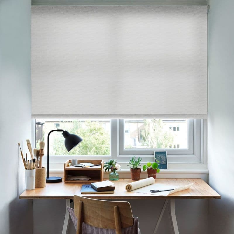 White fabric roller blind lowered nearly fully across the window, textured with a slim bottom gap; filtering soft daylight over a desk with lamp, plants and stationery, trees visible outside.