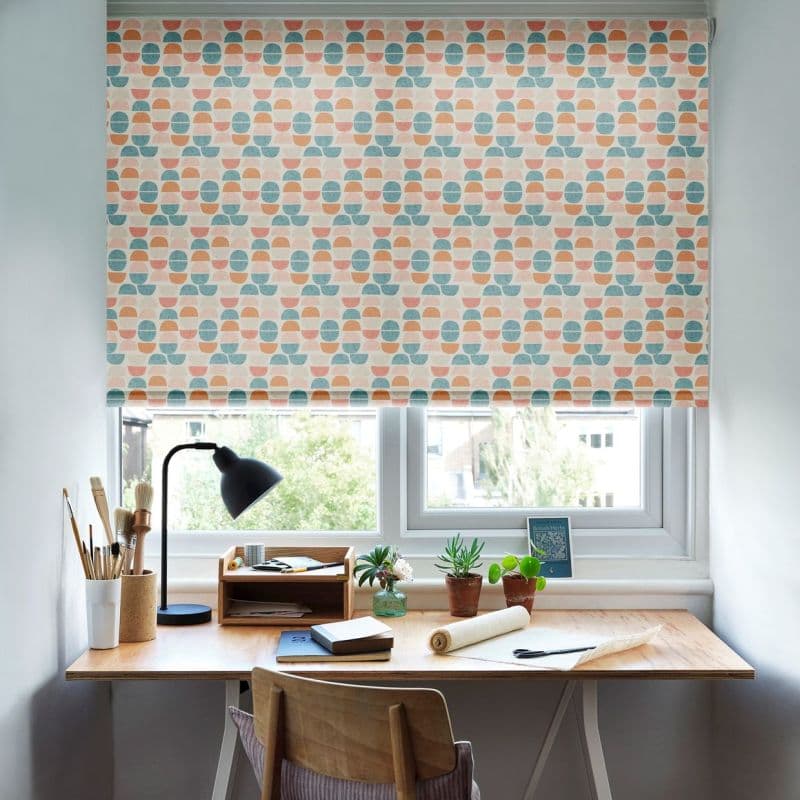 Patterned roller blind, lowered, featuring repeating teal, peach and terracotta semi‑circle motif; filtering soft daylight over a small home desk with lamp, art supplies, potted plants and a chair.