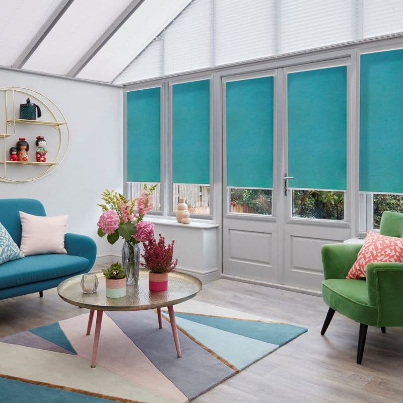 Teal roller blinds pulled down across tall conservatory windows, blocking view and filtering daylight with short clear strips at bottoms; sunroom furnished with colorful sofas, geometric rug, and coffee table.