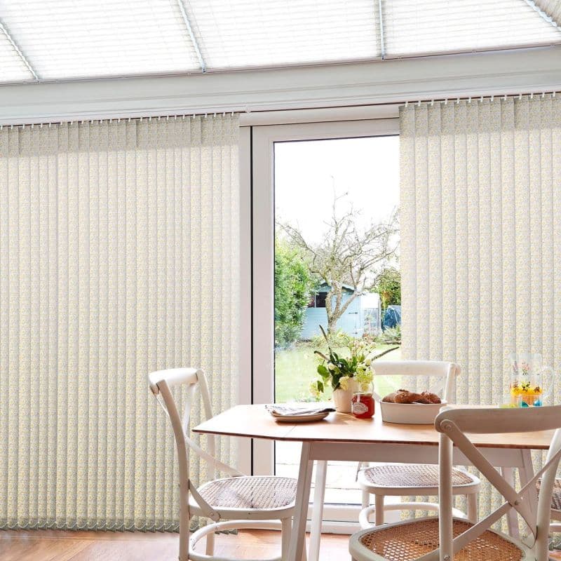 Light-patterned vertical fabric blinds hang mostly drawn, parted at the center to reveal a glass patio door; they filter daylight into a dining area with table, chairs and garden view.