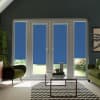 French doors and tall windows are covered by blue roller blinds, blocking most light; living room features green armchairs, glass coffee table on chevron rug, garden glimpses below.
